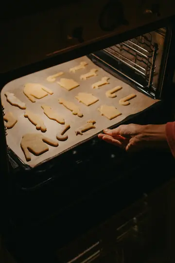 Detailní záběr ukazuje ruku, jak vkládá plech s různými vánočními sušenkami do teplé, tmavé trouby.