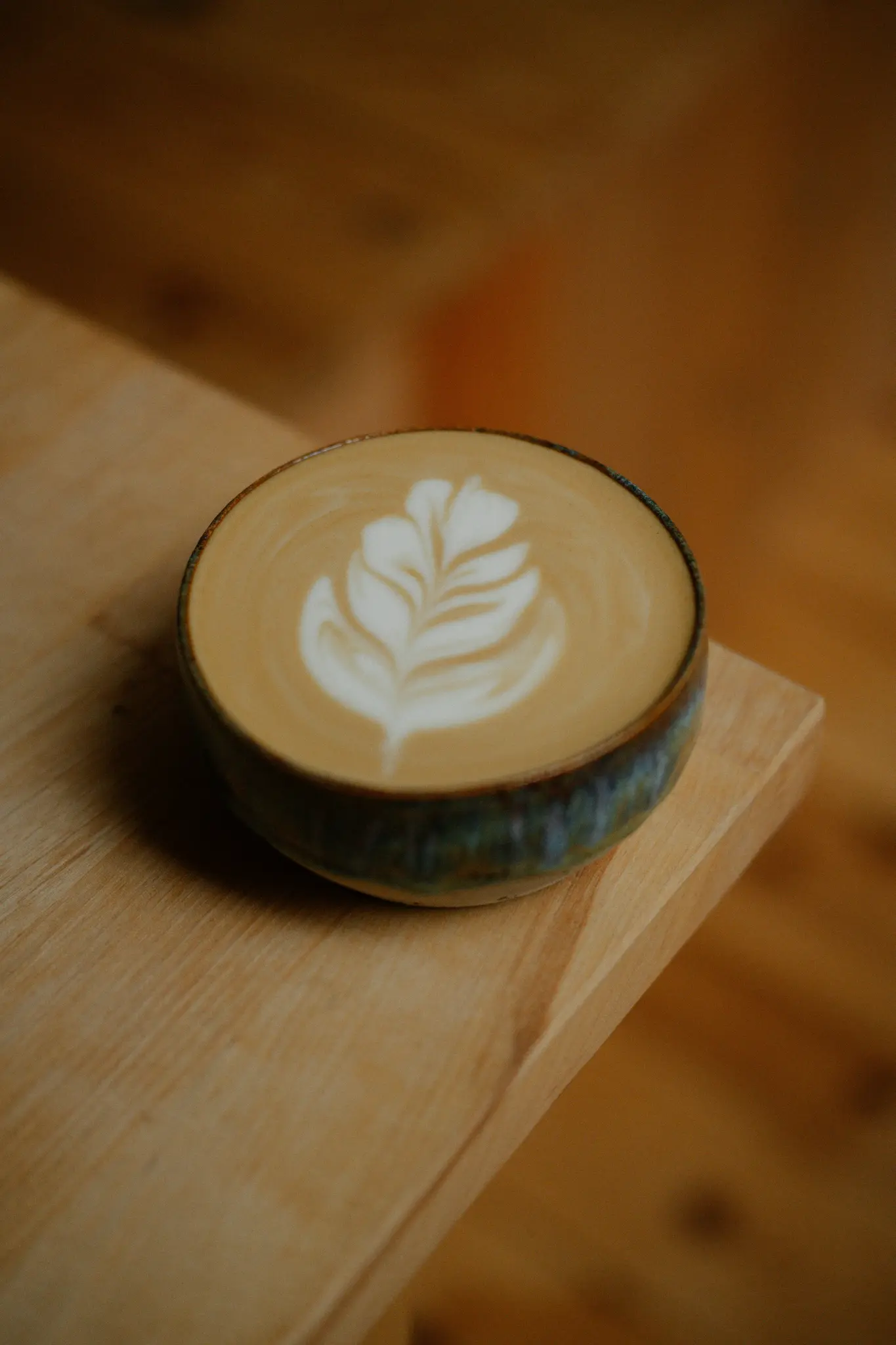 Detailní záběr latte v tmavě tyrkysovém keramickém šálku s detailním bílým listovým vzorem v pěně, položený na…