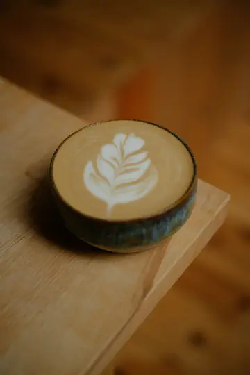 Detailní záběr latte v tmavě tyrkysovém keramickém šálku s detailním bílým listovým vzorem v pěně, položený na…