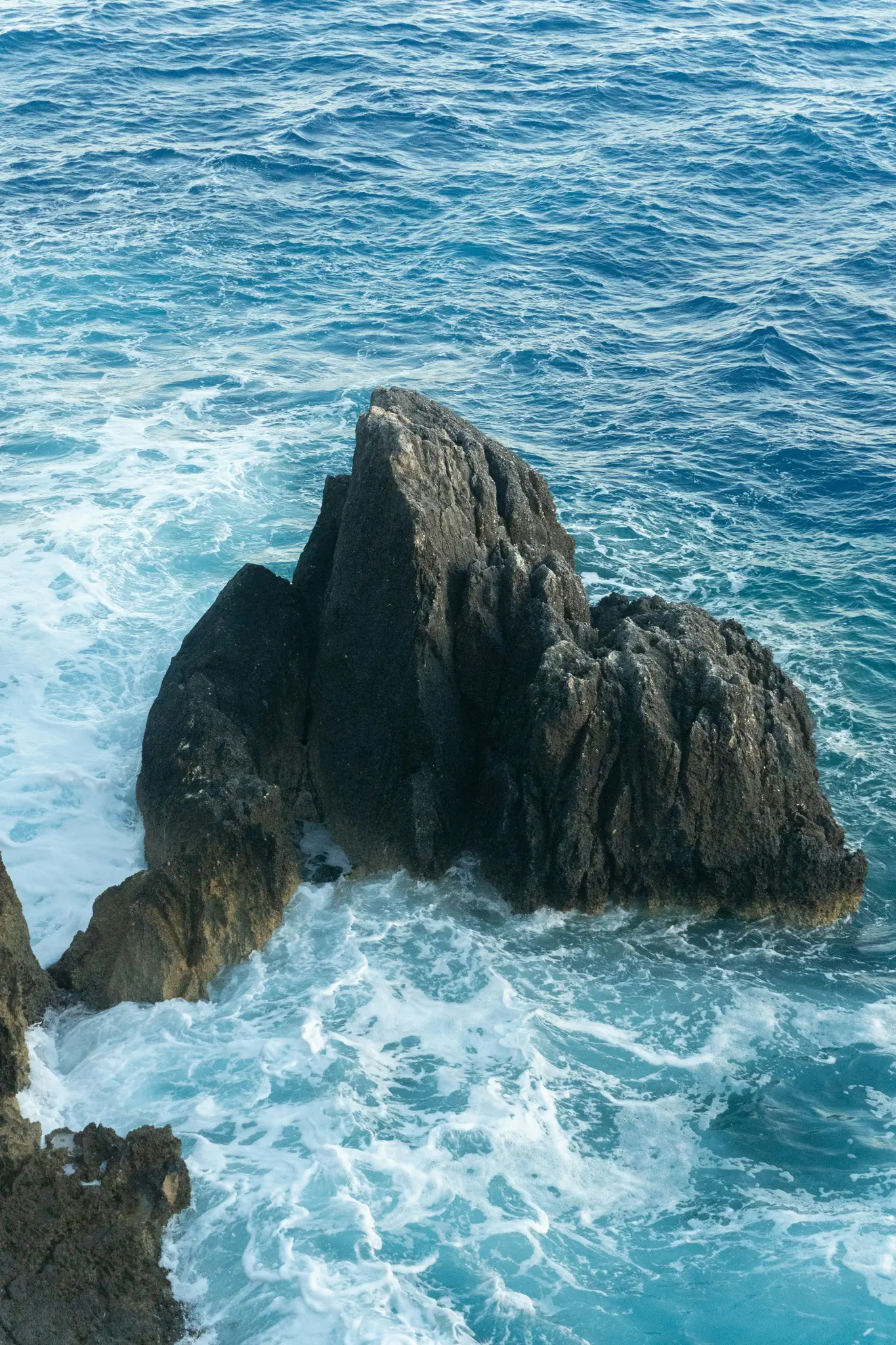 Eine Ansicht von oben zeigt dunkle, zerklüftete Felsen, die aus lebhaft blauem Ozeanwasser auftauchen, mit weißen, schaumigen