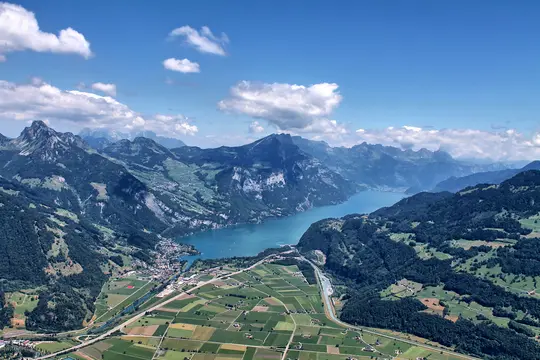 Et luftfoto viser en lang, smal, blå sø, der snor sig gennem en dal, flankeret af stejle, grønne bjerge under en blå himmel.