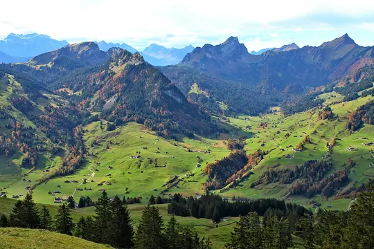 A panoramic view of a lush green valley dotted with small houses and winding paths, surrounded by mountains with autumn…