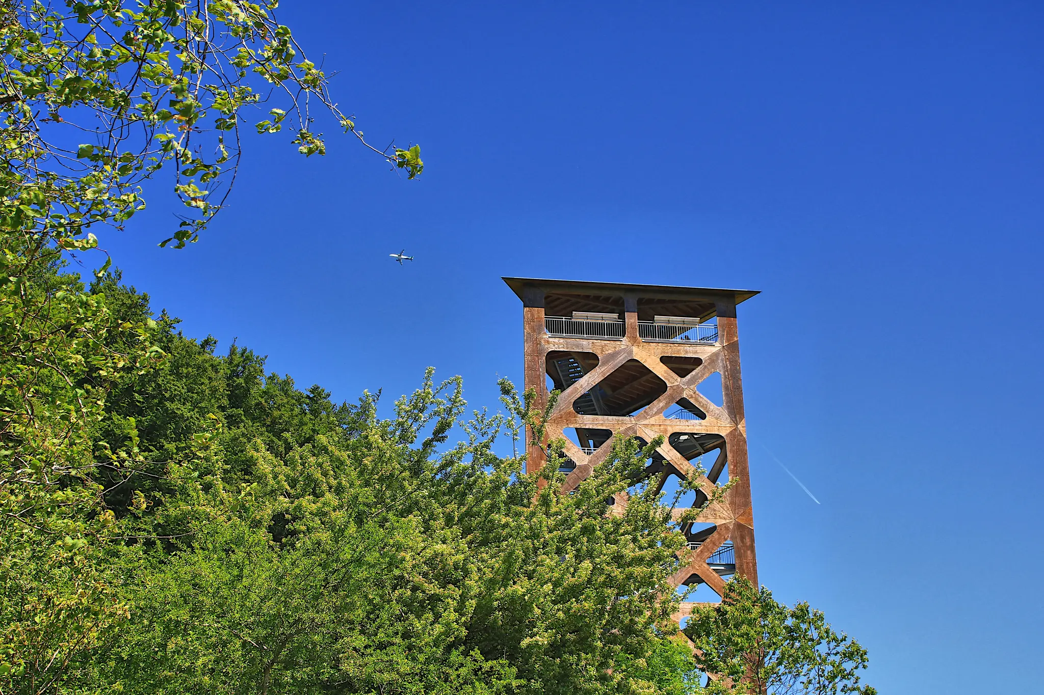 A rust-colored observation tower with a geometric design rises above green trees against a bright blue sky, with a small…