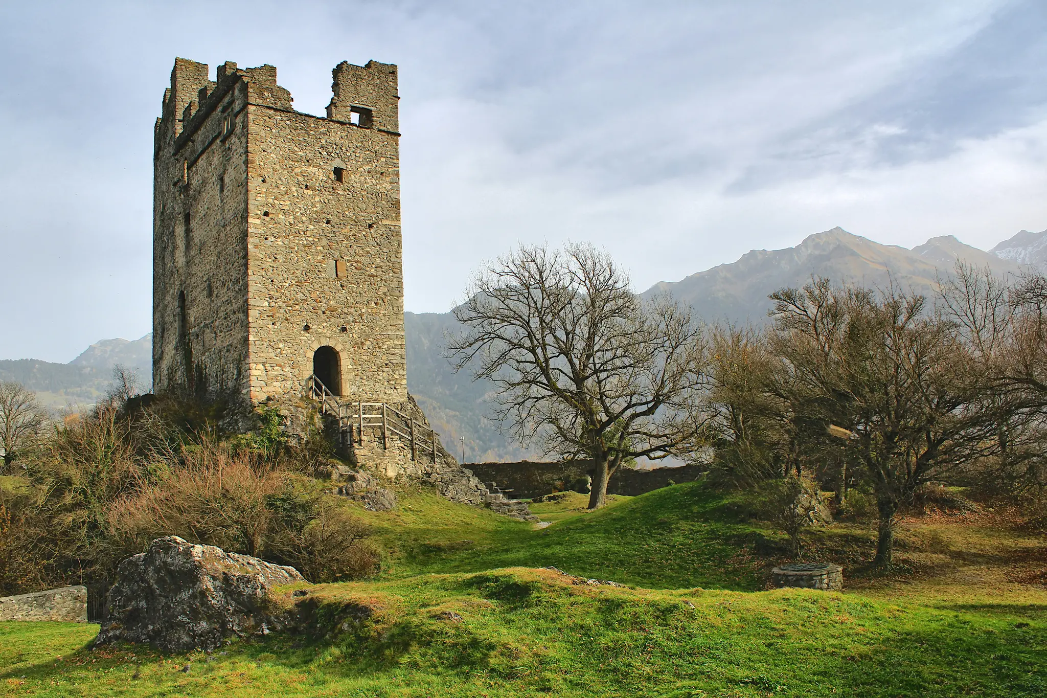 A tall, weathered stone tower with battlements stands on a green hill. Bare trees are scattered around, and mountains rise…