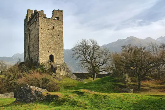 A tall, weathered stone tower with battlements stands on a green hill. Bare trees are scattered around, and mountains rise…