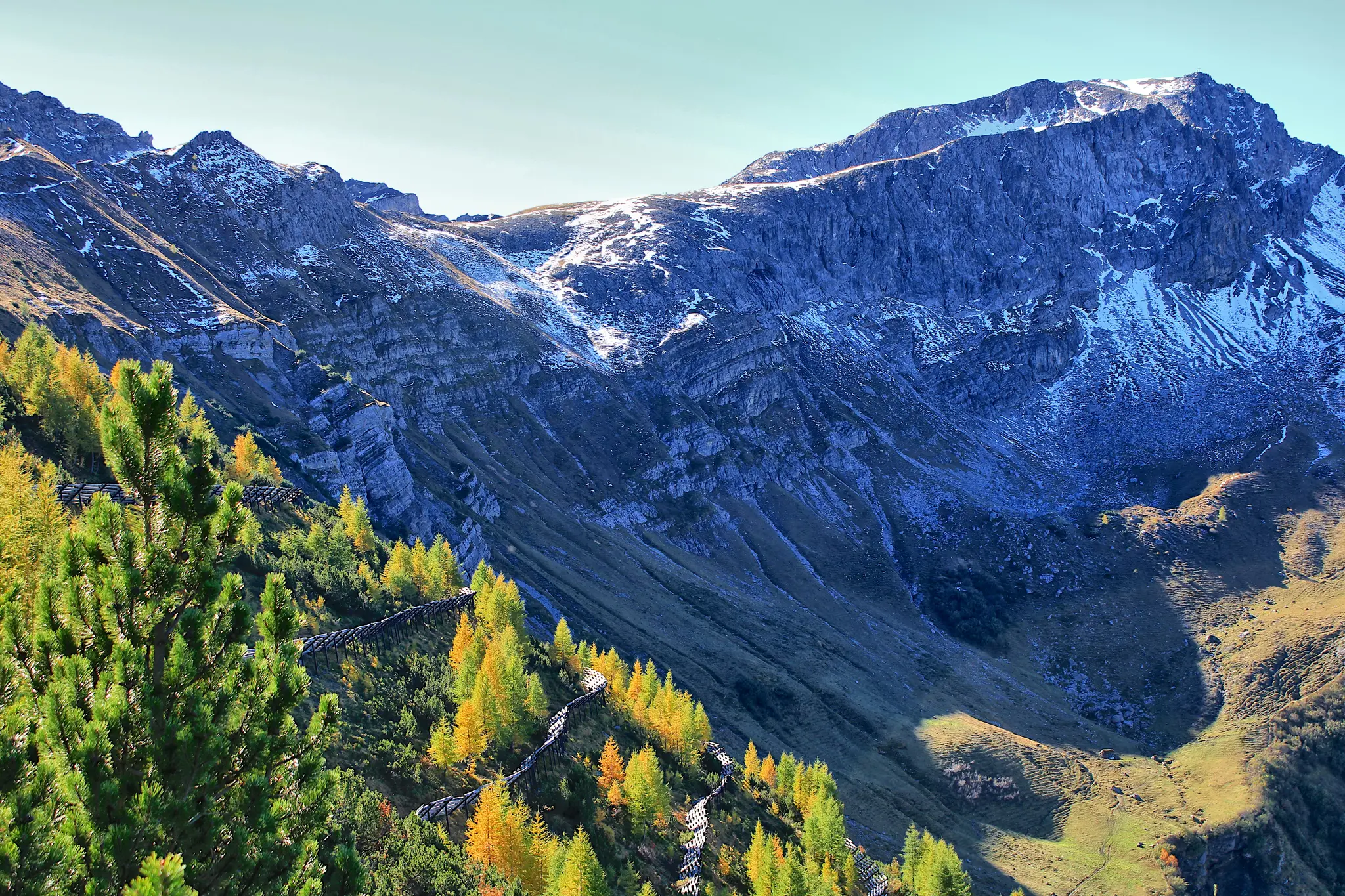 A wide landscape view of a mountain range with a prominent snow-dusted peak under a clear sky. The foreground features a…