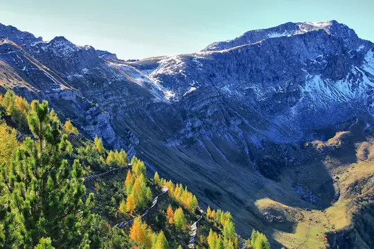 A wide landscape view of a mountain range with a prominent snow-dusted peak under a clear sky. The foreground features a…