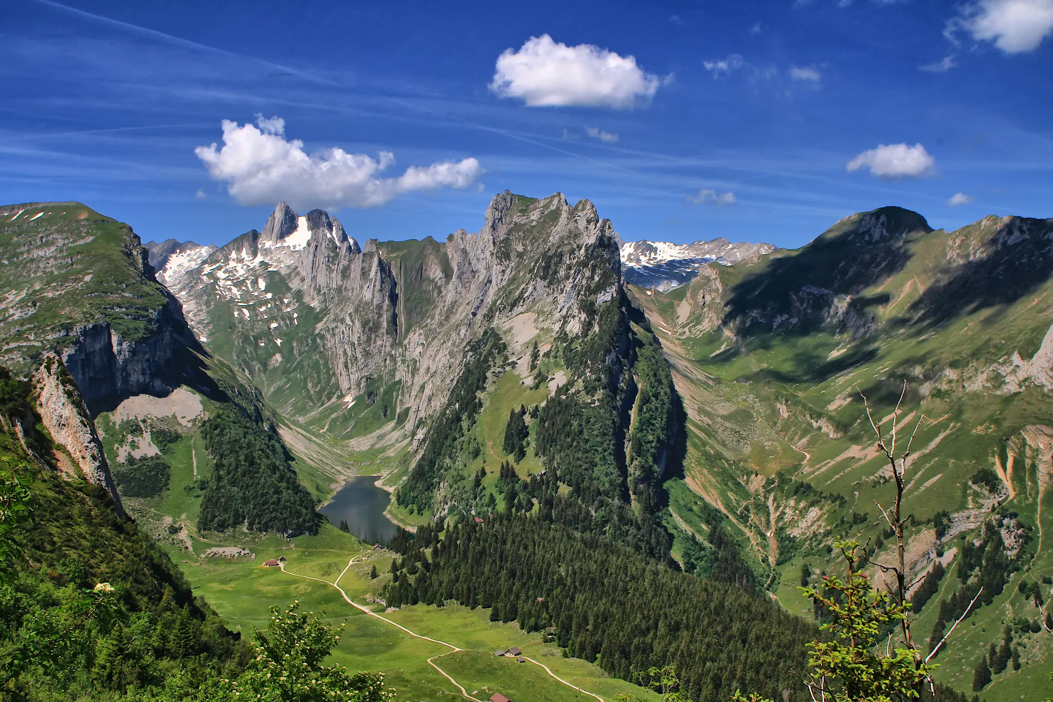 An aerial view of a vast mountain landscape under a blue sky with white clouds. A dark lake is visible in a green valley.