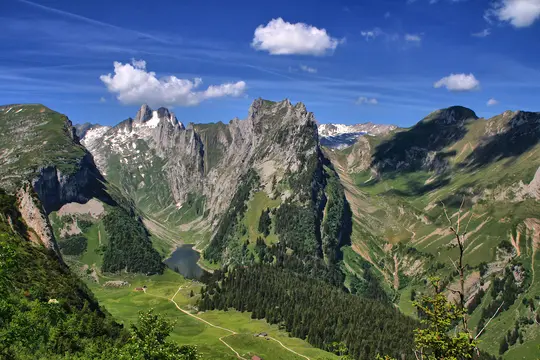 An aerial view of a vast mountain landscape under a blue sky with white clouds. A dark lake is visible in a green valley.