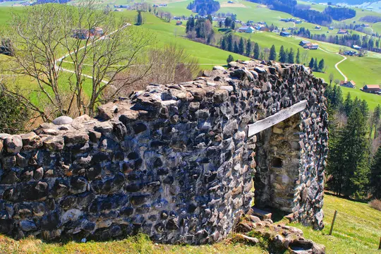 Et nærbillede af gamle stenruiner i forgrunden, med en levende grøn dal, snoede veje og spredte huse i baggrunden.