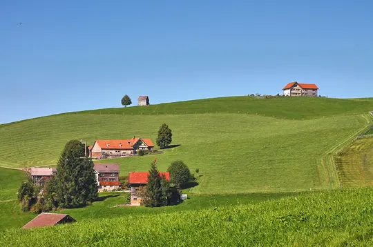 A wide landscape view of rolling green hills under a clear blue sky. Several houses with red roofs are nestled among trees…