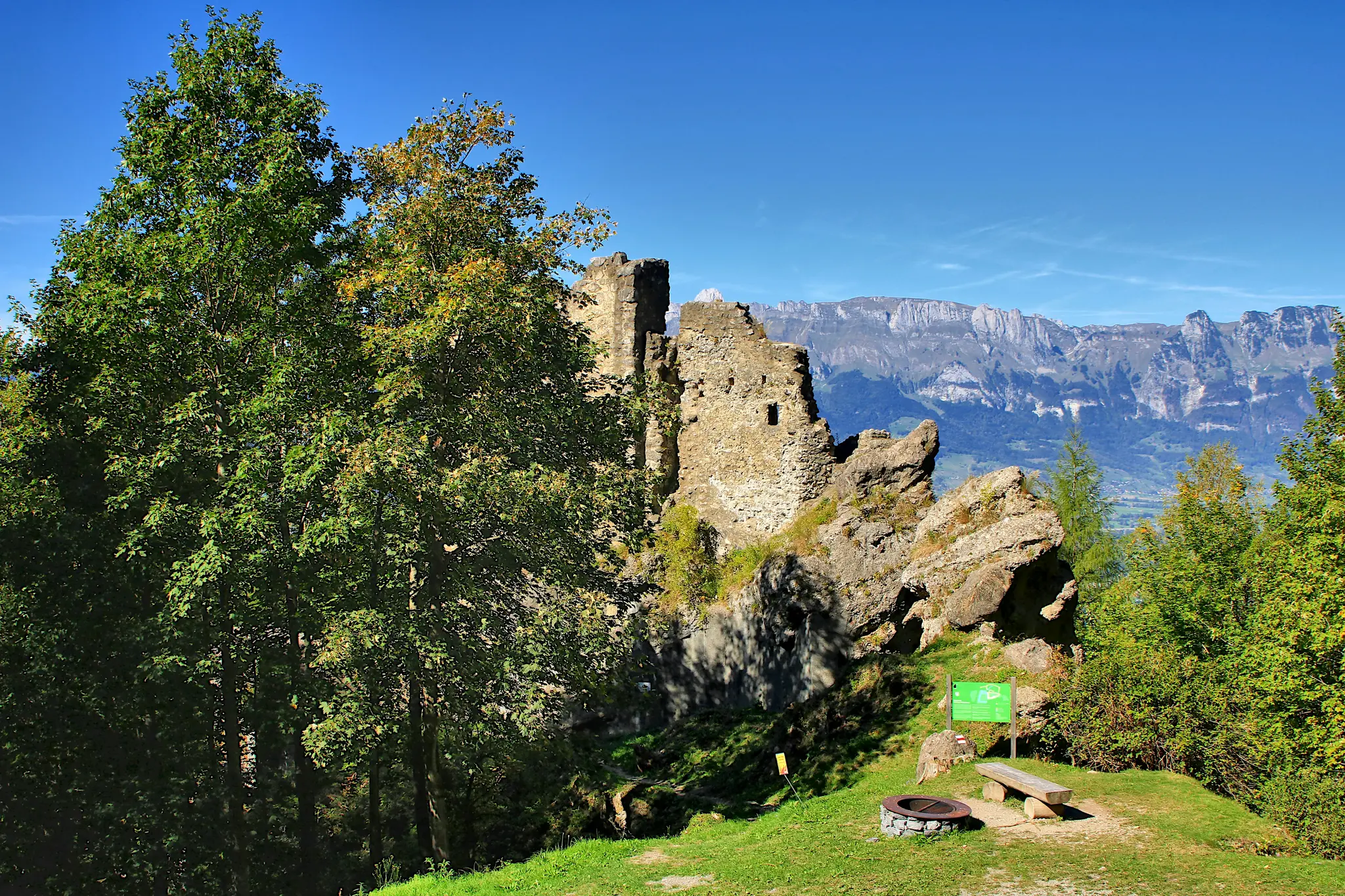 A wide landscape view of ancient stone castle ruins on a grassy hill, flanked by green and slightly yellowing trees. In the…