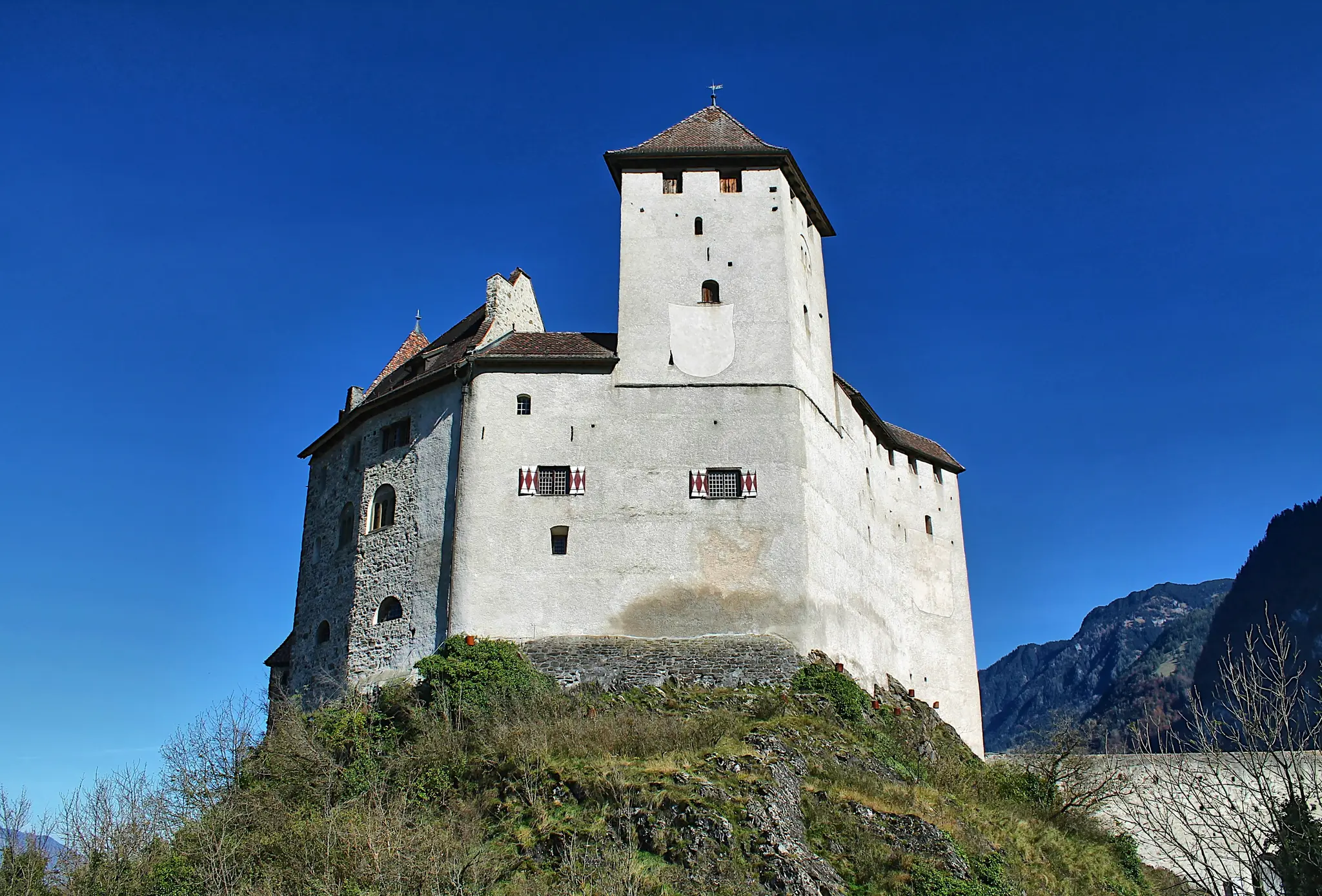 A large, light-colored castle with a tall central tower and red-shuttered windows sits atop a rocky, green hill against a…