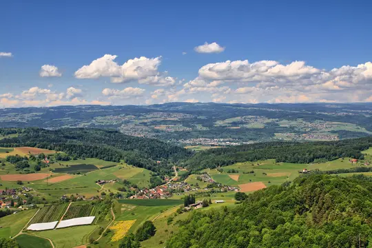 A wide panoramic shot of a lush green landscape featuring rolling hills, dense forests, agricultural fields, and small…