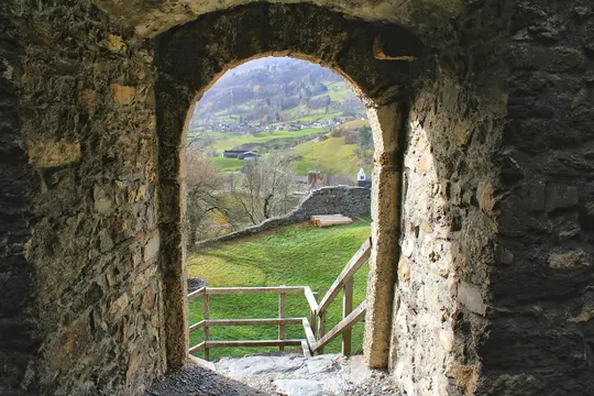 A view through a rough stone archway reveals a vibrant green valley with rolling hills, scattered buildings, and bare trees.