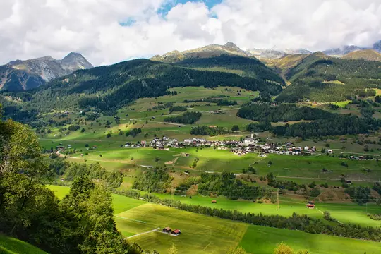 A panoramic view of a small village with white and brown roofs in a lush green valley, surrounded by forested mountains…
