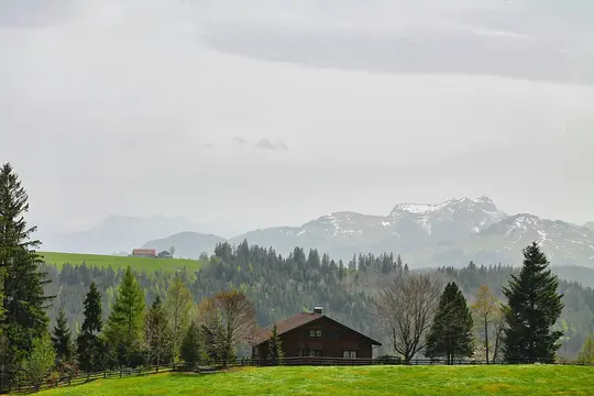 Et brunt hus med rødt tag på en klar grøn bakke, omgivet af forskellige træer, med disede, snedækkede bjerge i baggrunden.