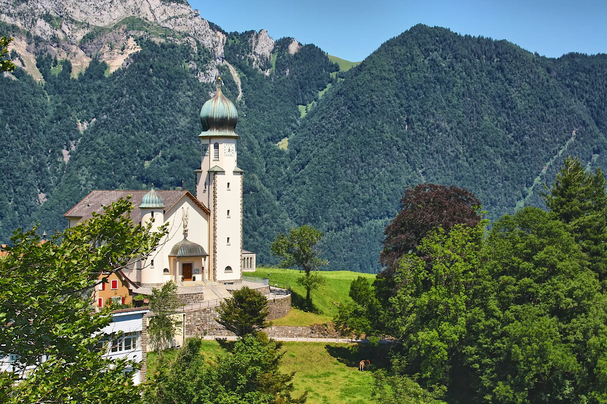 A white church with a green domed roof and tall bell tower stands on a green hill, surrounded by trees and mountains.