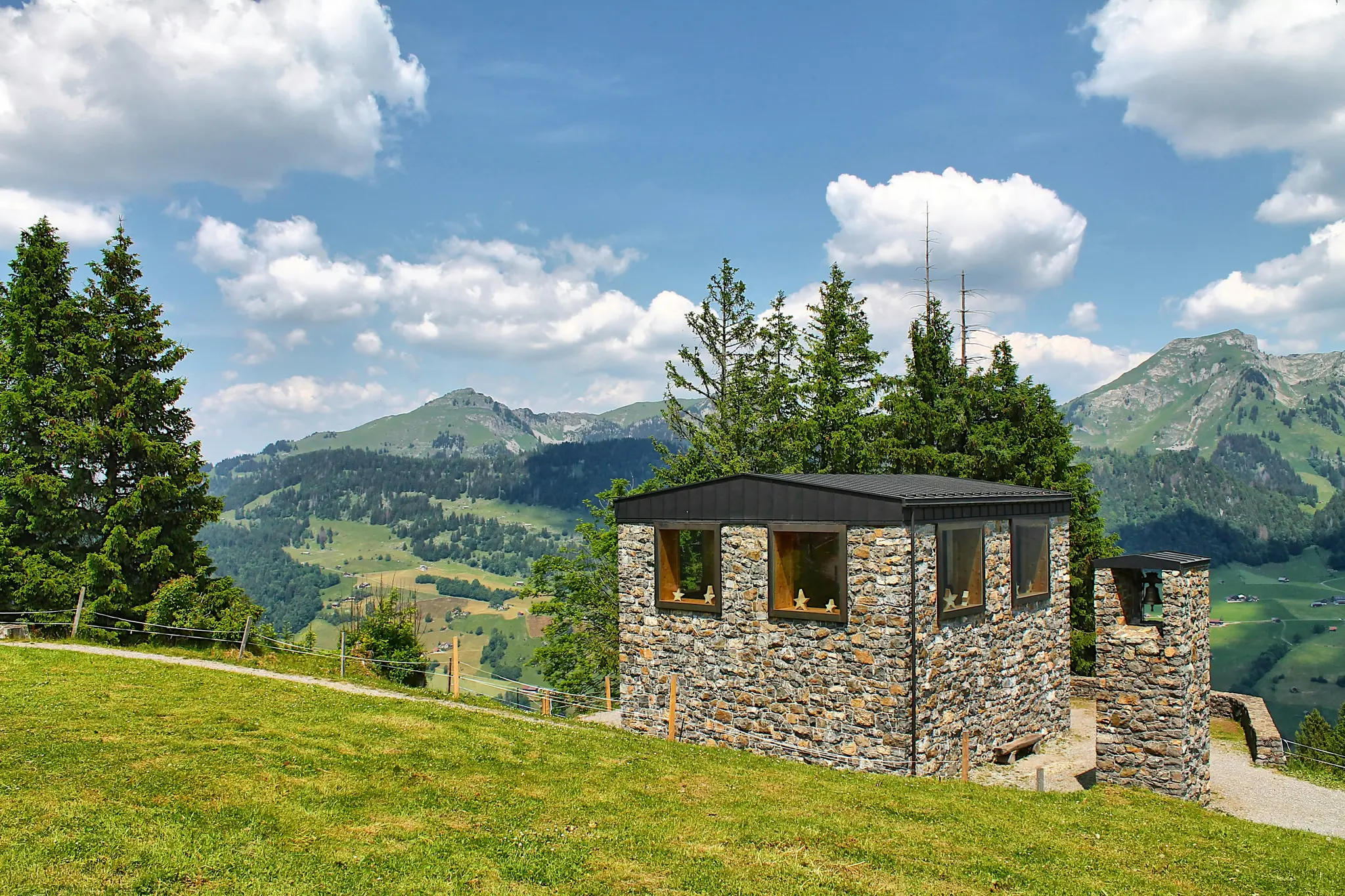 A stone building with a black roof and wooden windows sits on a vibrant green hill, overlooking a vast mountain range and…
