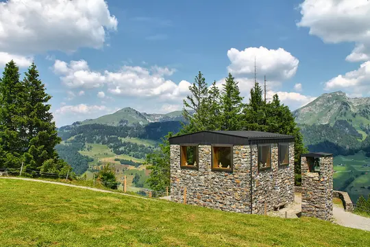 A stone building with a black roof and wooden windows sits on a vibrant green hill, overlooking a vast mountain range and…