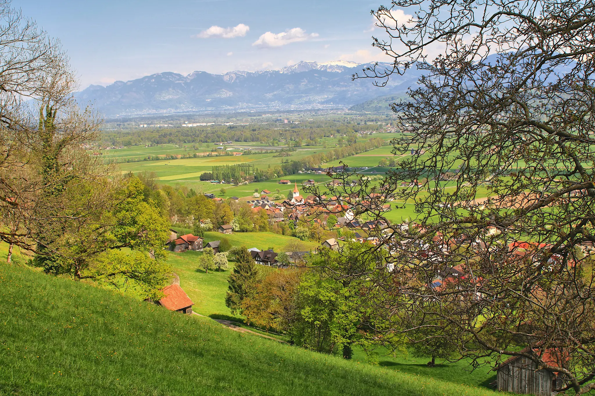 An elevated wide landscape view of a lush green valley with a small village nestled among trees, backed by snow-capped…