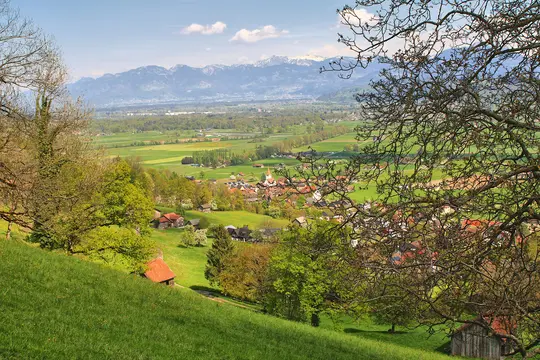An elevated wide landscape view of a lush green valley with a small village nestled among trees, backed by snow-capped…