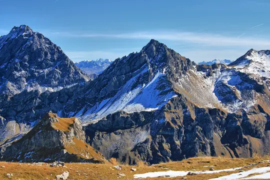A wide landscape photo of a rugged mountain range. Peaks are dark grey and rocky, with streaks and patches of white snow.…