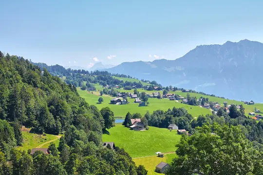 A wide shot of a verdant mountain valley with numerous small houses scattered across bright green fields. A small pond is…