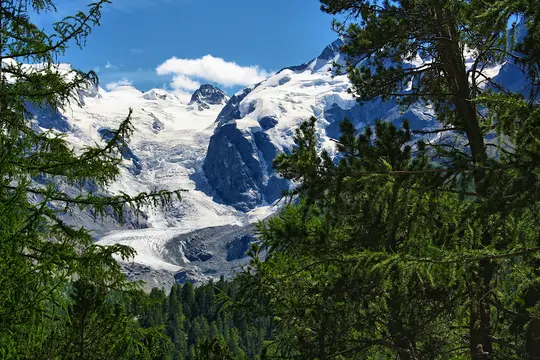 A wide landscape photo showing a vast glacier flowing down from snow-capped mountains under a blue sky, framed by green…