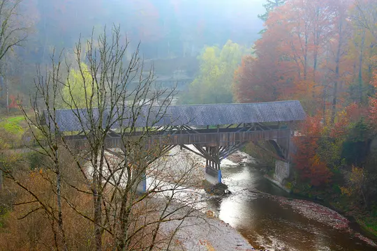 A wooden covered bridge with a shingled roof crosses a shallow river. Bare trees are in the foreground, and hillsides with…