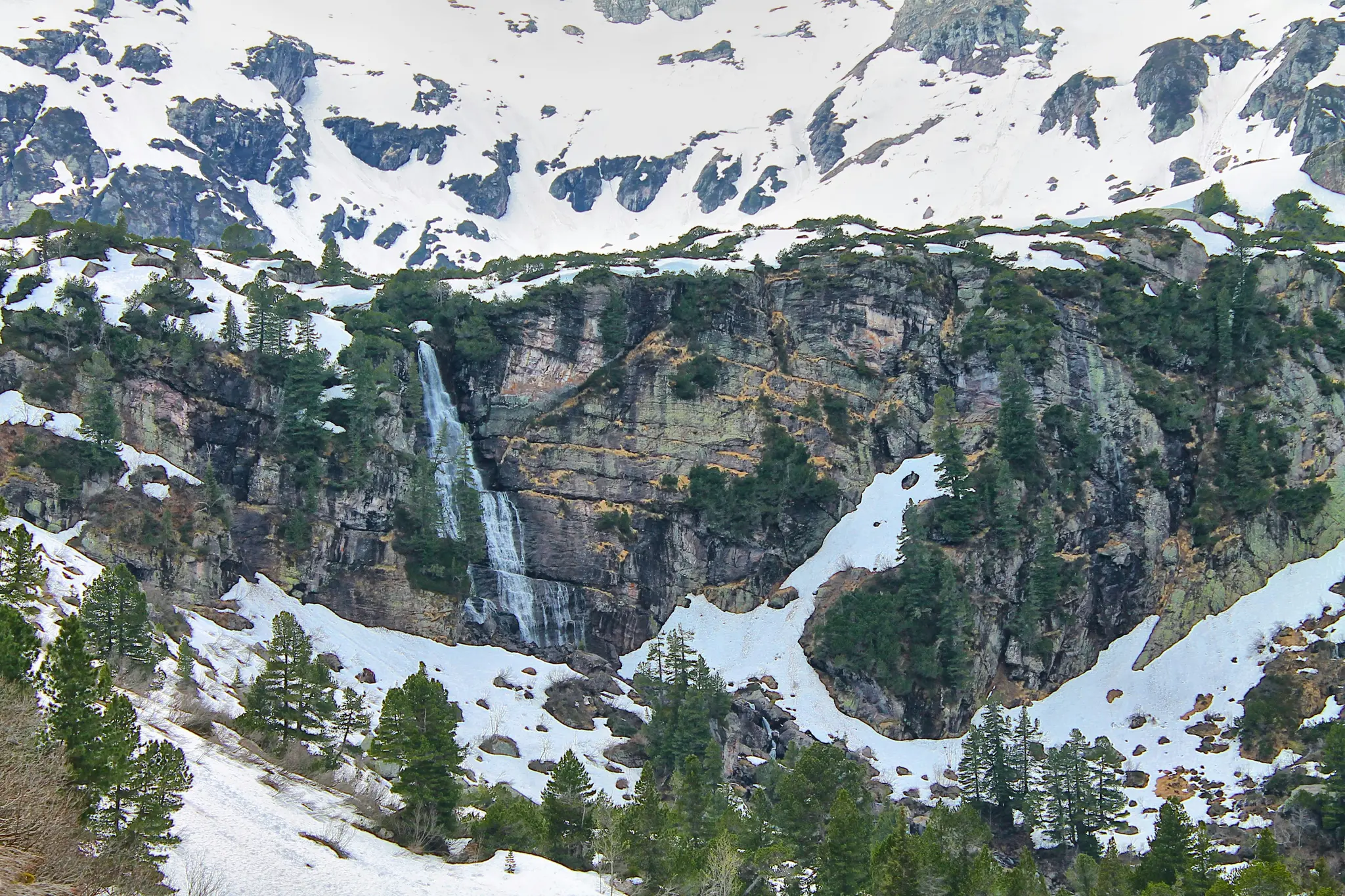 A tall waterfall flows down a dark, rugged mountain cliff. Patches of white snow and green evergreen trees dot the rocky…