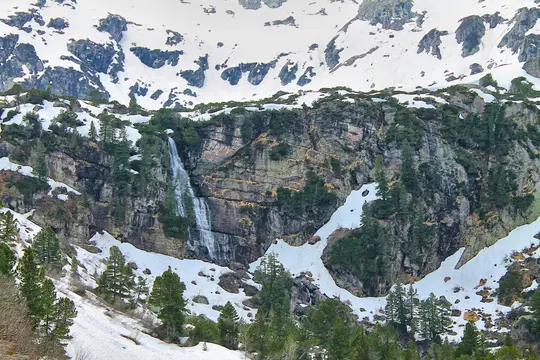 A tall waterfall flows down a dark, rugged mountain cliff. Patches of white snow and green evergreen trees dot the rocky…
