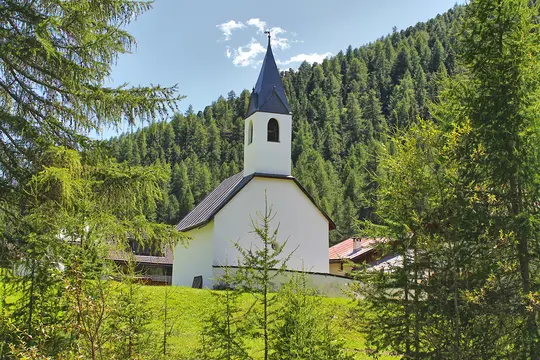 En lille hvid kirke med et mørkegråt spir midt i levende grønne træer, med en tæt skov der dækker en bakketop…