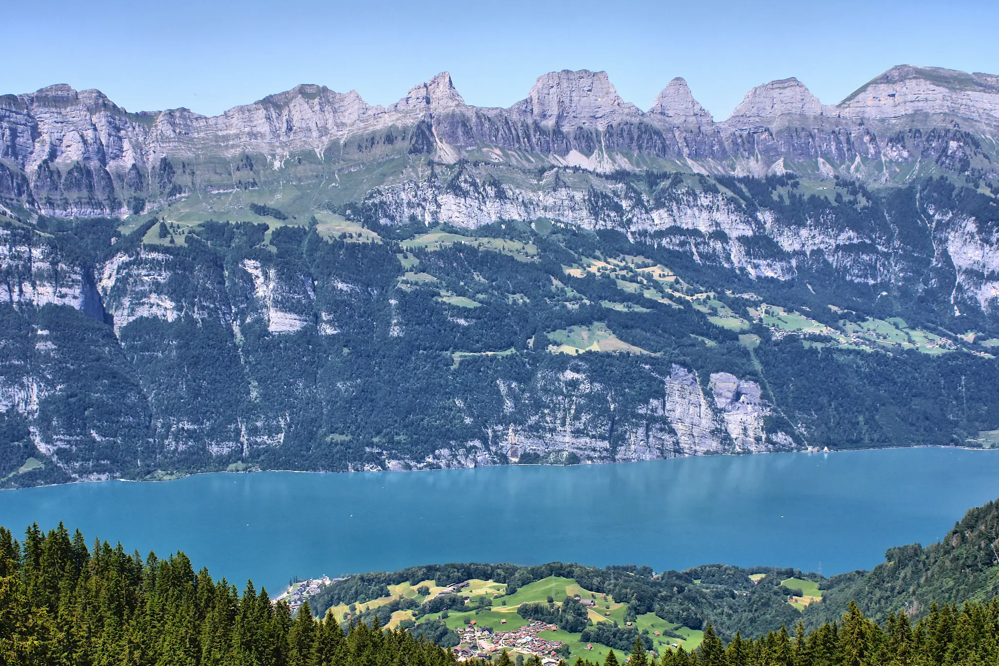 A panoramic view of a bright blue lake surrounded by lush green forests and steep, rocky mountains under a clear sky.