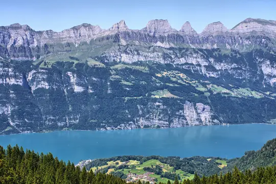 A panoramic view of a bright blue lake surrounded by lush green forests and steep, rocky mountains under a clear sky.