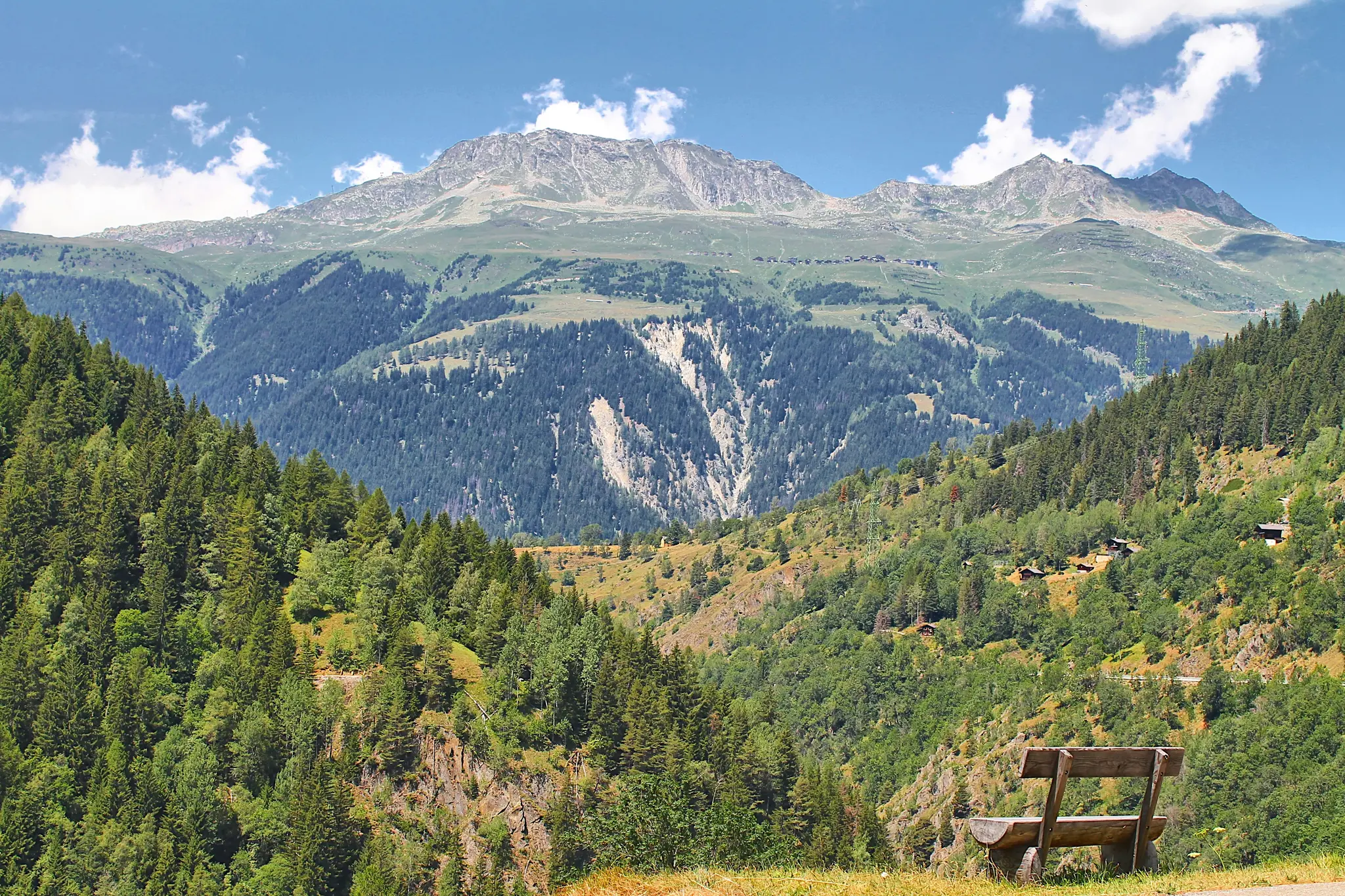 A rustic wooden bench sits on a grassy hill, facing a wide mountain valley filled with lush green forests and distant peaks.