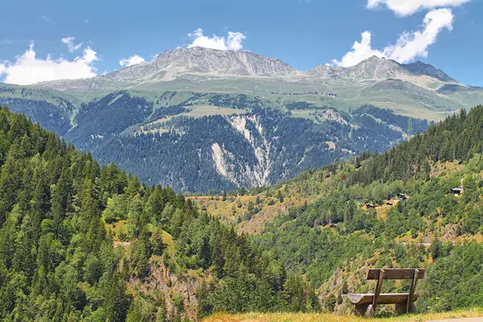 A rustic wooden bench sits on a grassy hill, facing a wide mountain valley filled with lush green forests and distant peaks.