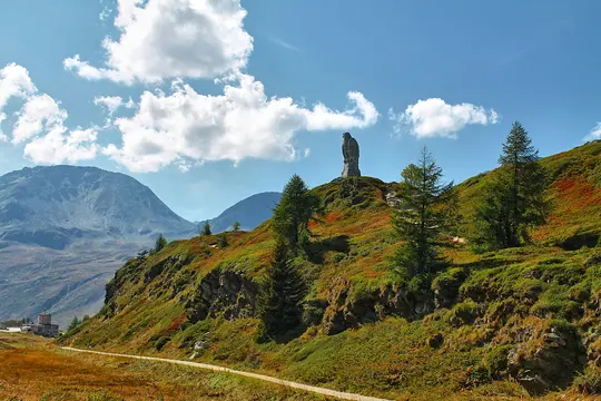 A large stone eagle statue overlooks a vast mountainous landscape with a winding path and sparse trees under a bright blue…