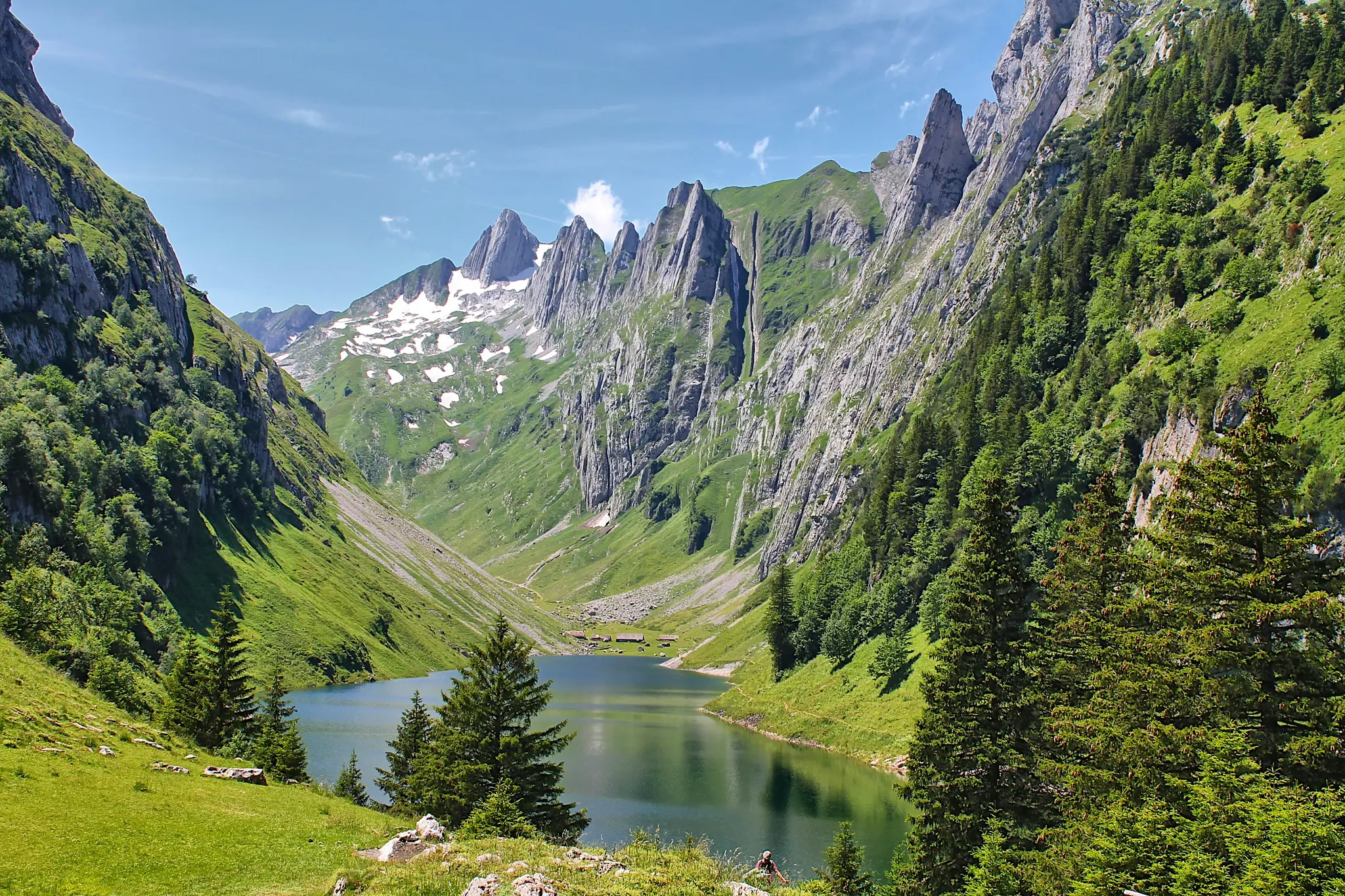 A wide landscape photo shows a calm, dark blue lake in a deep valley, flanked by steep green mountains and jagged rocky…
