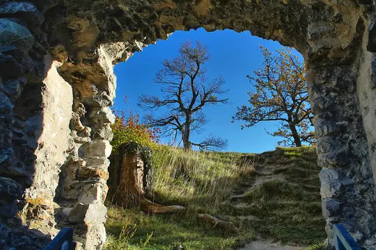 A view through a rough stone archway reveals a grassy hill under a bright blue sky. A bare tree stands prominently on the…