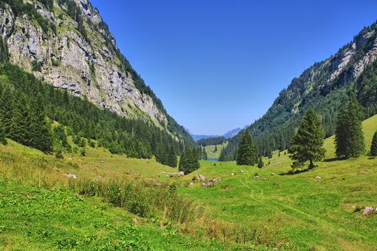 A wide landscape photo of a verdant mountain valley. Steep, rocky mountainsides covered in dense green trees frame a bright…