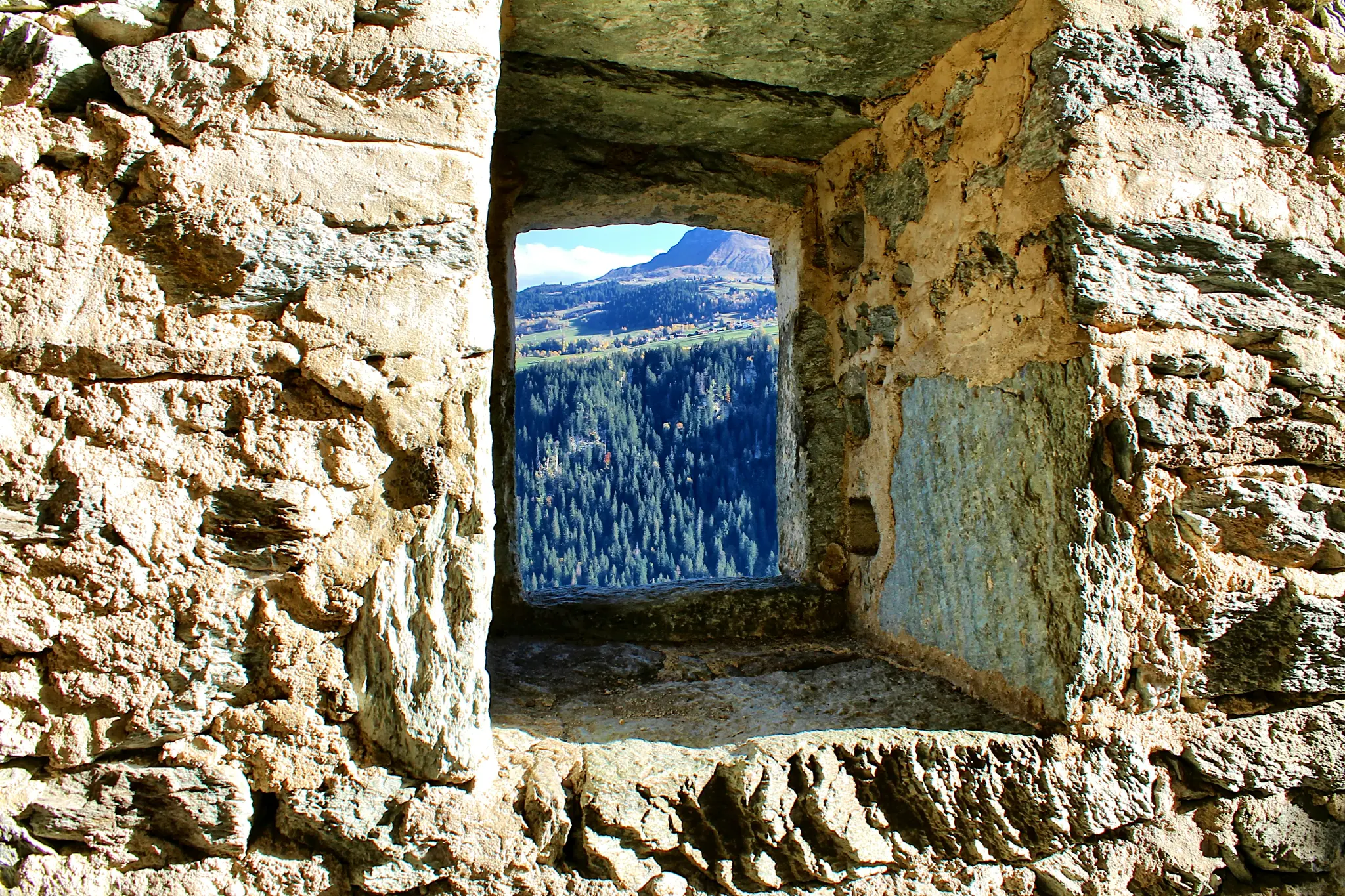 A rustic stone window frames a vibrant autumn mountain landscape. A dense green forest fills the midground, with a village…