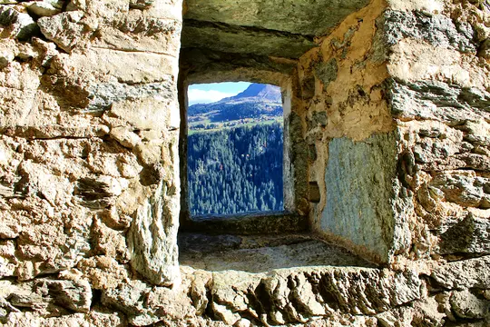 A rustic stone window frames a vibrant autumn mountain landscape. A dense green forest fills the midground, with a village…