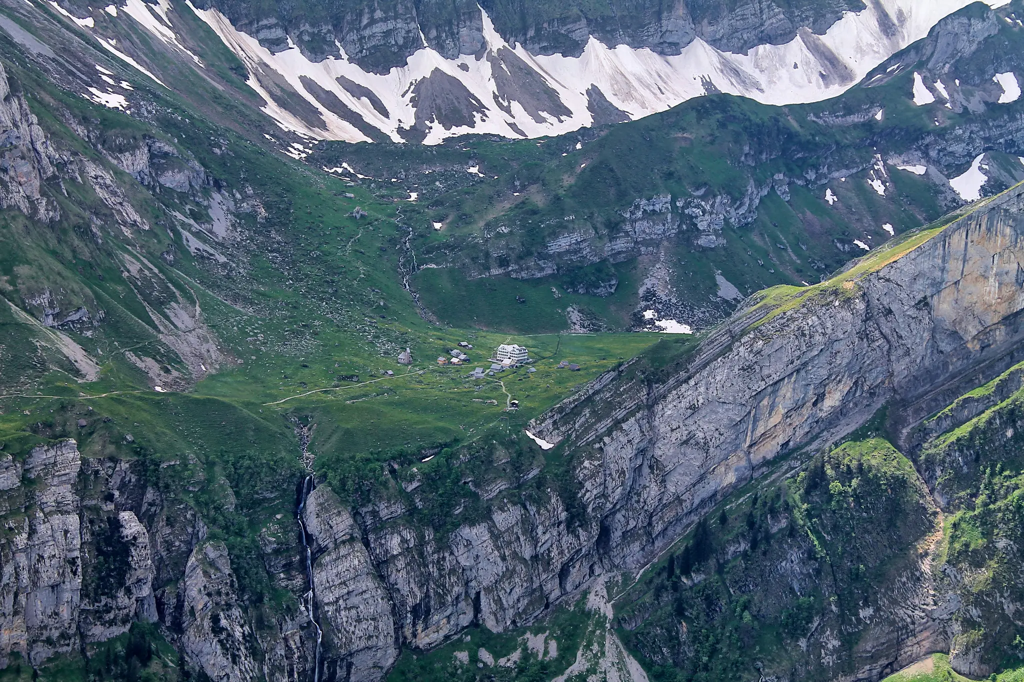 An aerial view shows a lush green alpine valley with a small village of buildings nestled among steep, rocky mountainsides…