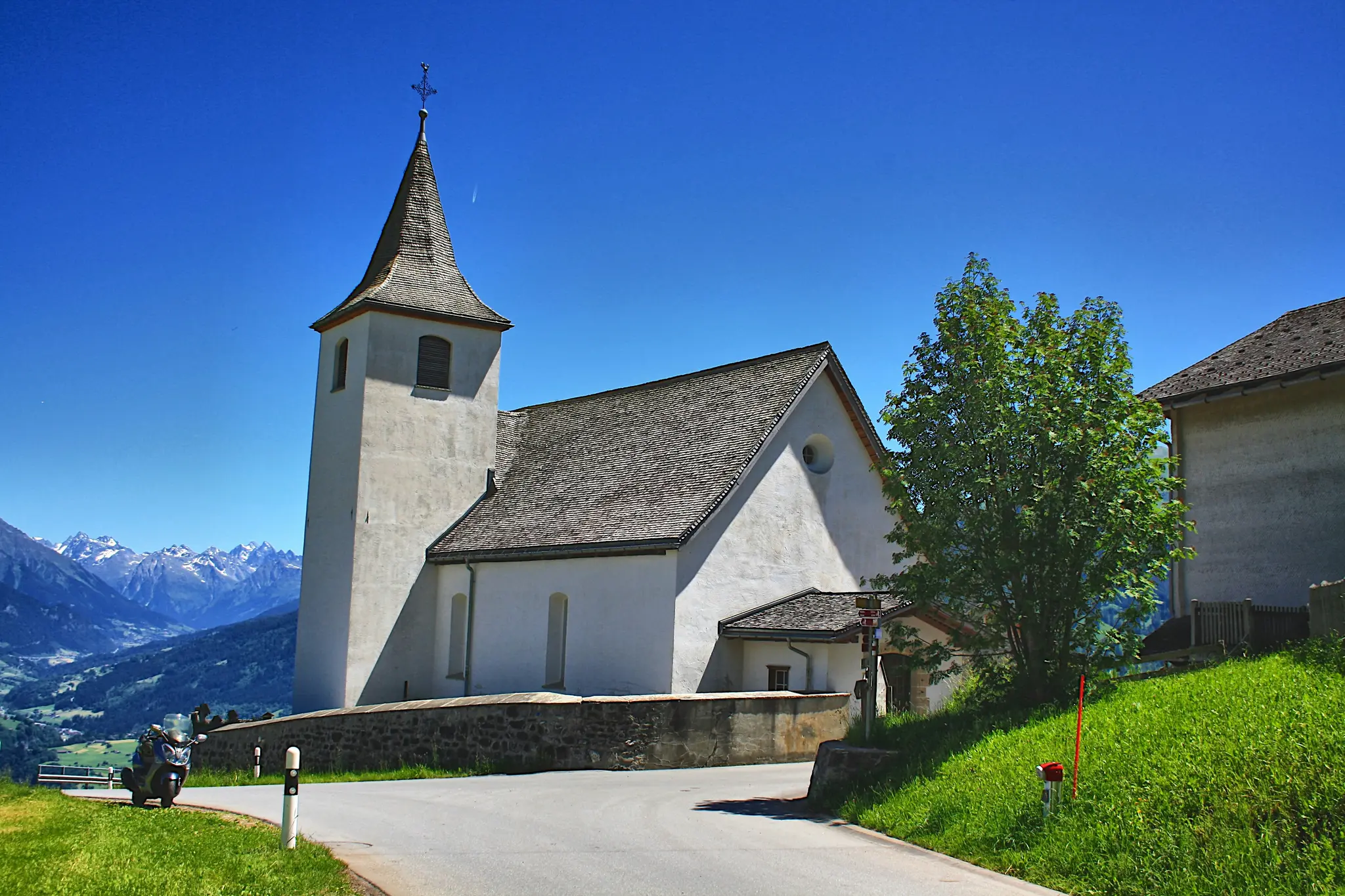 A white church with a tall steeple and a cross on top stands on a green hillside. A winding road is in the foreground.…