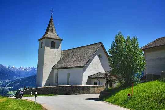 A white church with a tall steeple and a cross on top stands on a green hillside. A winding road is in the foreground.…
