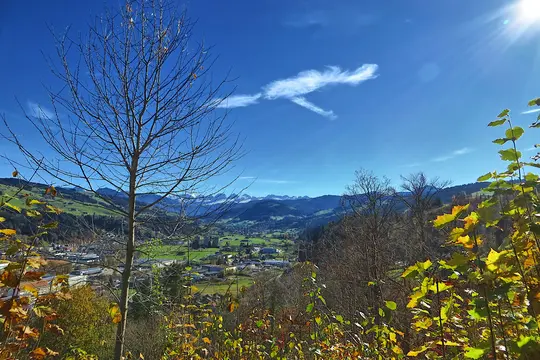 A vibrant autumn landscape shows a valley town nestled among green fields and hills, with distant snow-capped mountains.