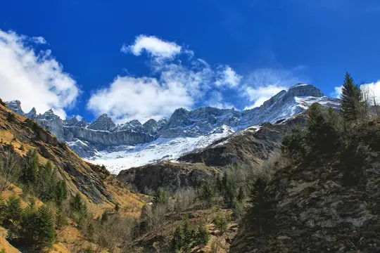 En panoramaudsigt over en barsk bjergkæde med sne, der dækker toppene og de øvre skråninger. Himlen er en levende blå med…