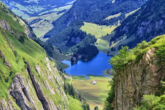 A high-angle view of a bright blue lake in a deep valley, flanked by steep, lush green mountains and forests.