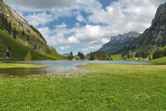 A wide shot of a tranquil mountain lake with lush green slopes on either side and a small village nestled by the far shore.